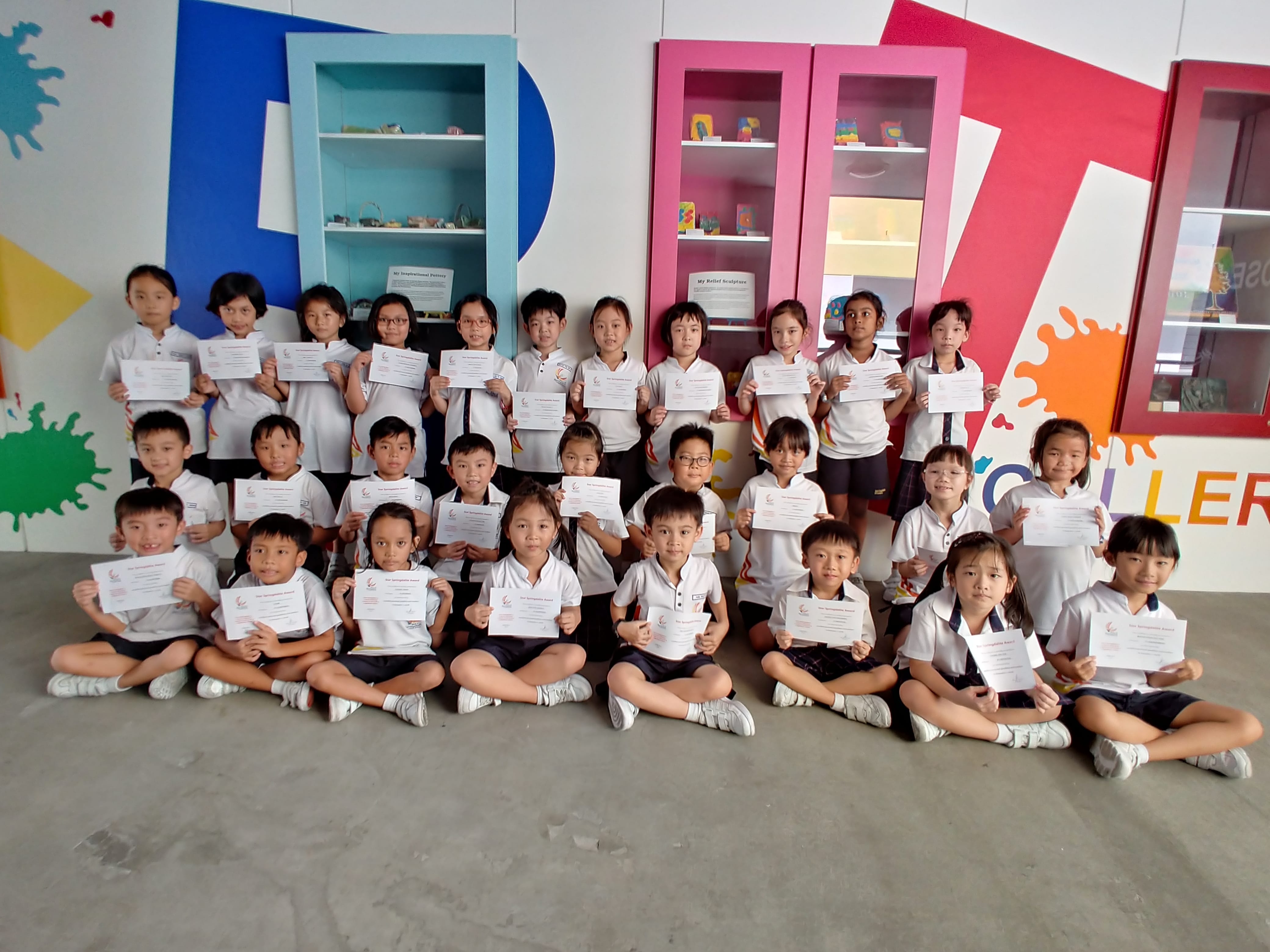 Group of young students in white uniforms holding certificates, posed in three rows in a colorful room.